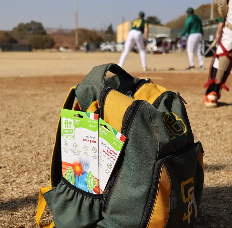 A green and gold sports backpack sits on a dirt baseball field with players blurred in the background. Two packs of FitPatch Therapy patches are displayed in the front pockets of the bag.