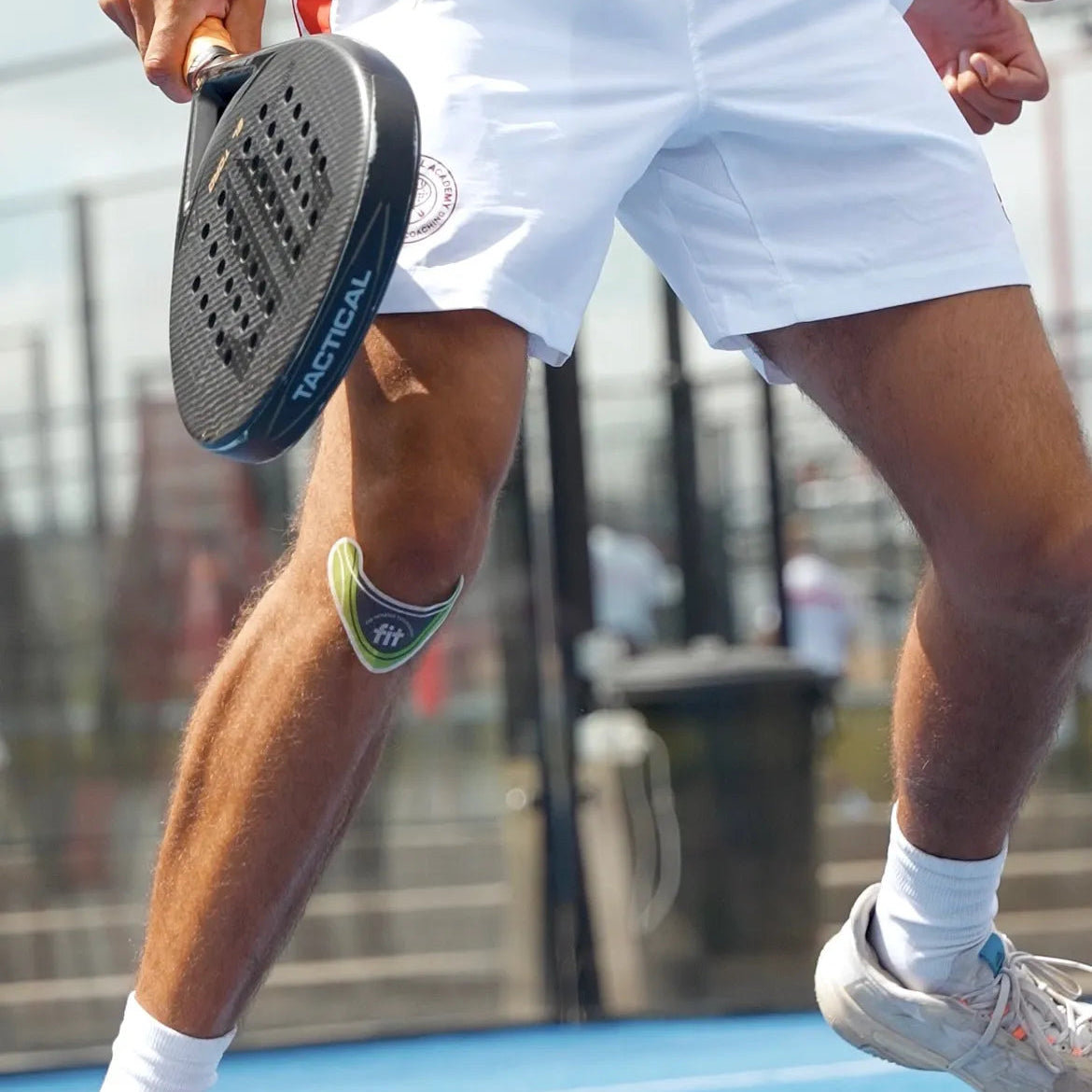 A close-up lifestyle shot of a padel player in white athletic wear holding a black RACTUS padel paddle, with a green and black FitPatch Therapy knee patch on their leg, on an outdoor court.