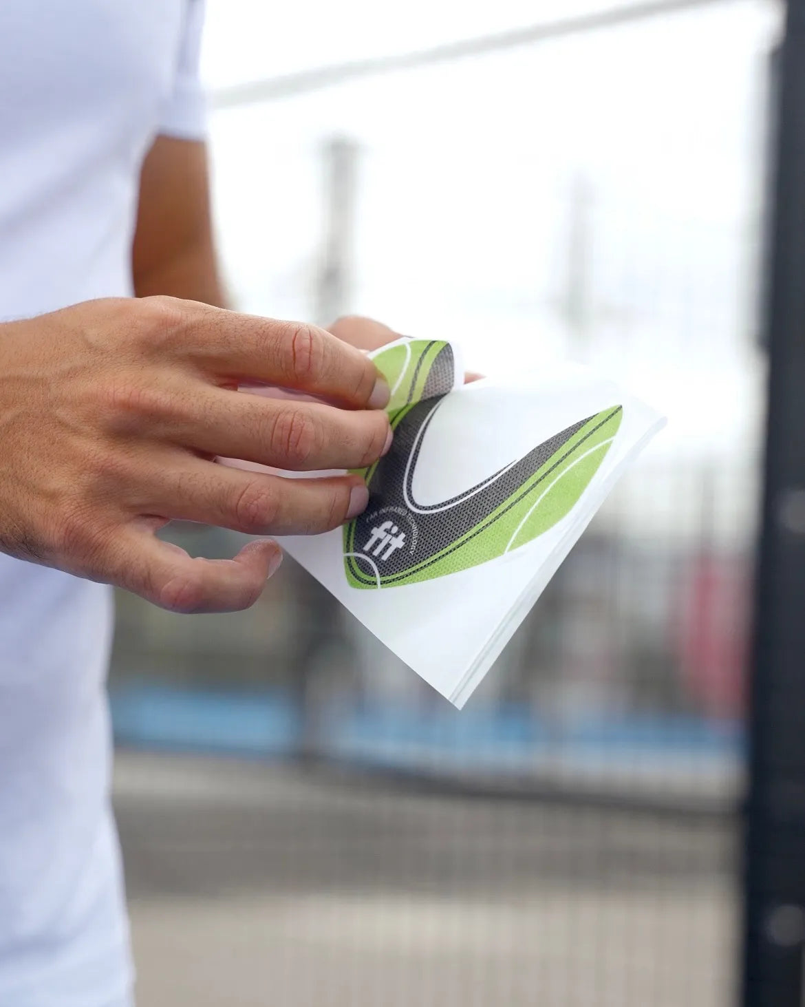 A close-up of a person's hand peeling a green, black, and white FitPatch Therapy Patch from a white backing sheet, with a blurred outdoor background.