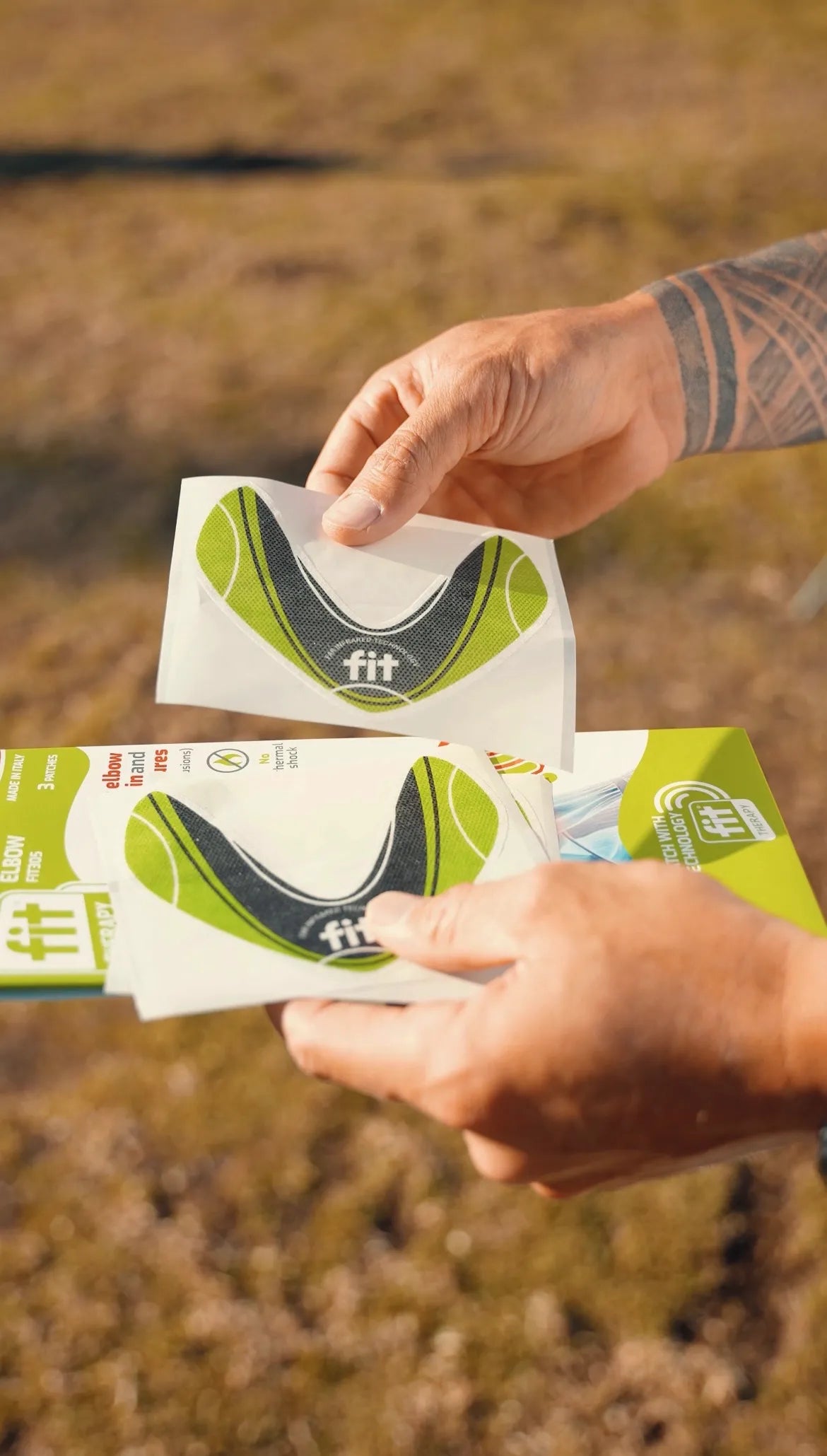 A close-up of a person's hands holding FitPatch Therapy patches with a green and black curved design, with a blurred outdoor grassy background.