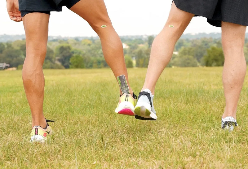 Three lower legs of people standing on a grassy field, wearing black athletic shorts and running shoes with small circular green FitPatch Therapy patches on their upper calves.
