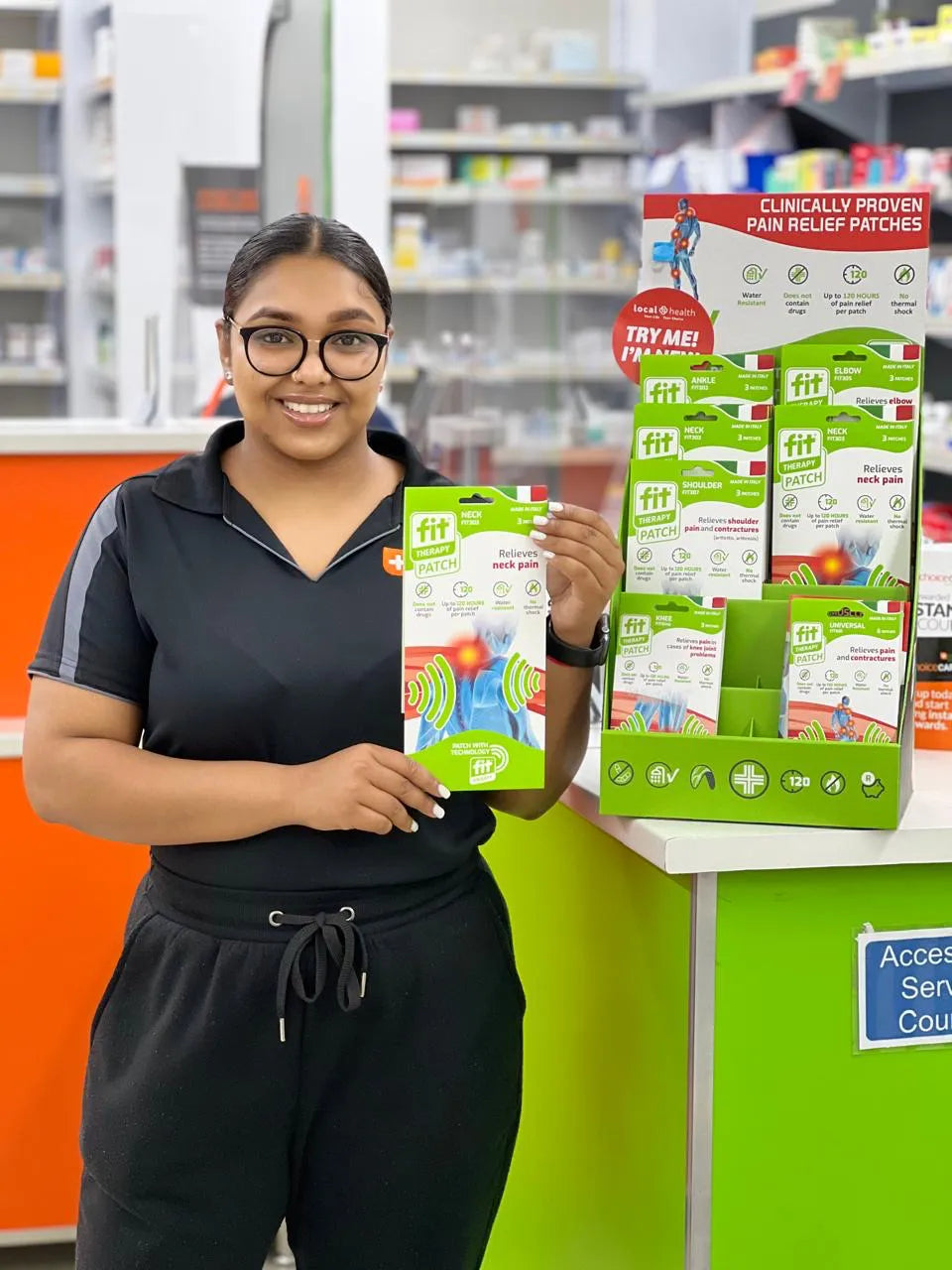 A woman in a black athletic polo shirt and sweatpants stands in a retail store holding a package of FitPatch Therapy pain relief patches, next to a green and white display stand of the same product.