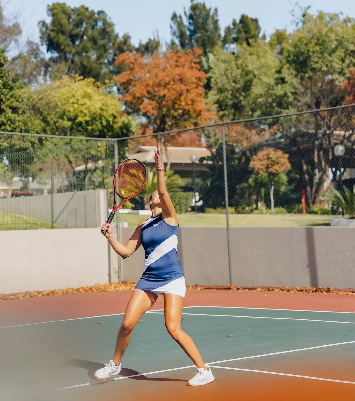 A woman playing tennis on an outdoor clay court, wearing a navy blue and white athletic dress and white sneakers, holding a tennis racket with both hands ready to hit the ball.